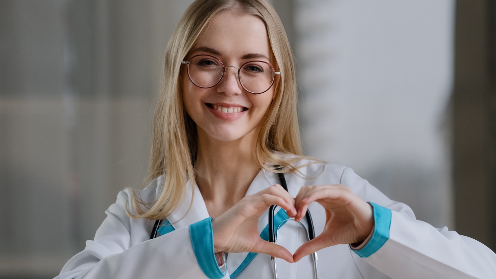 Młoda, uśmiechnięta kobieta w lekarskim kitlu składa z palców kształt serca