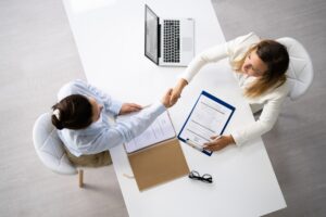 Widok z góry - biurko przy którym siedzą dwie kobiety. Podają sobie dłonie. Na biurku laptop i dokumenty